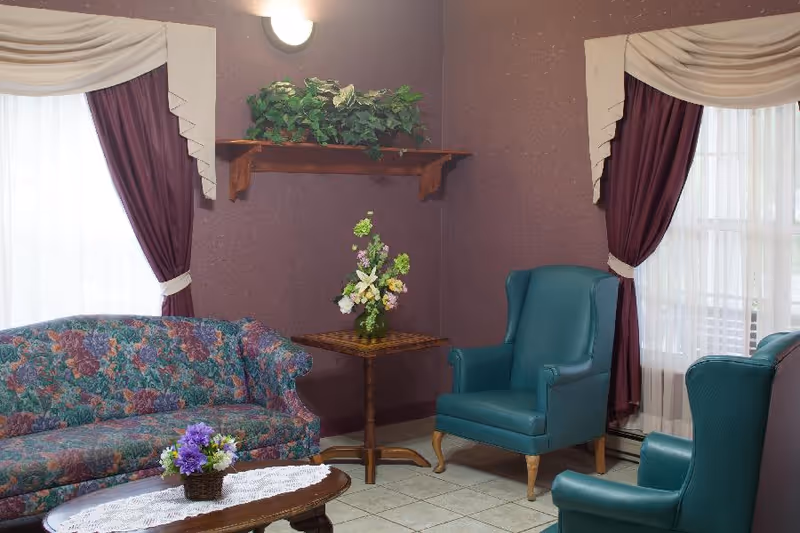 A cozy living room with a floral patterned sofa, two teal wingback chairs, a wooden coffee table with a lace doily and a small flower arrangement, a corner table with a vase of flowers, purple walls, two windows with sheer white curtains and purple drapes, and a wooden shelf with green plants above the corner table.