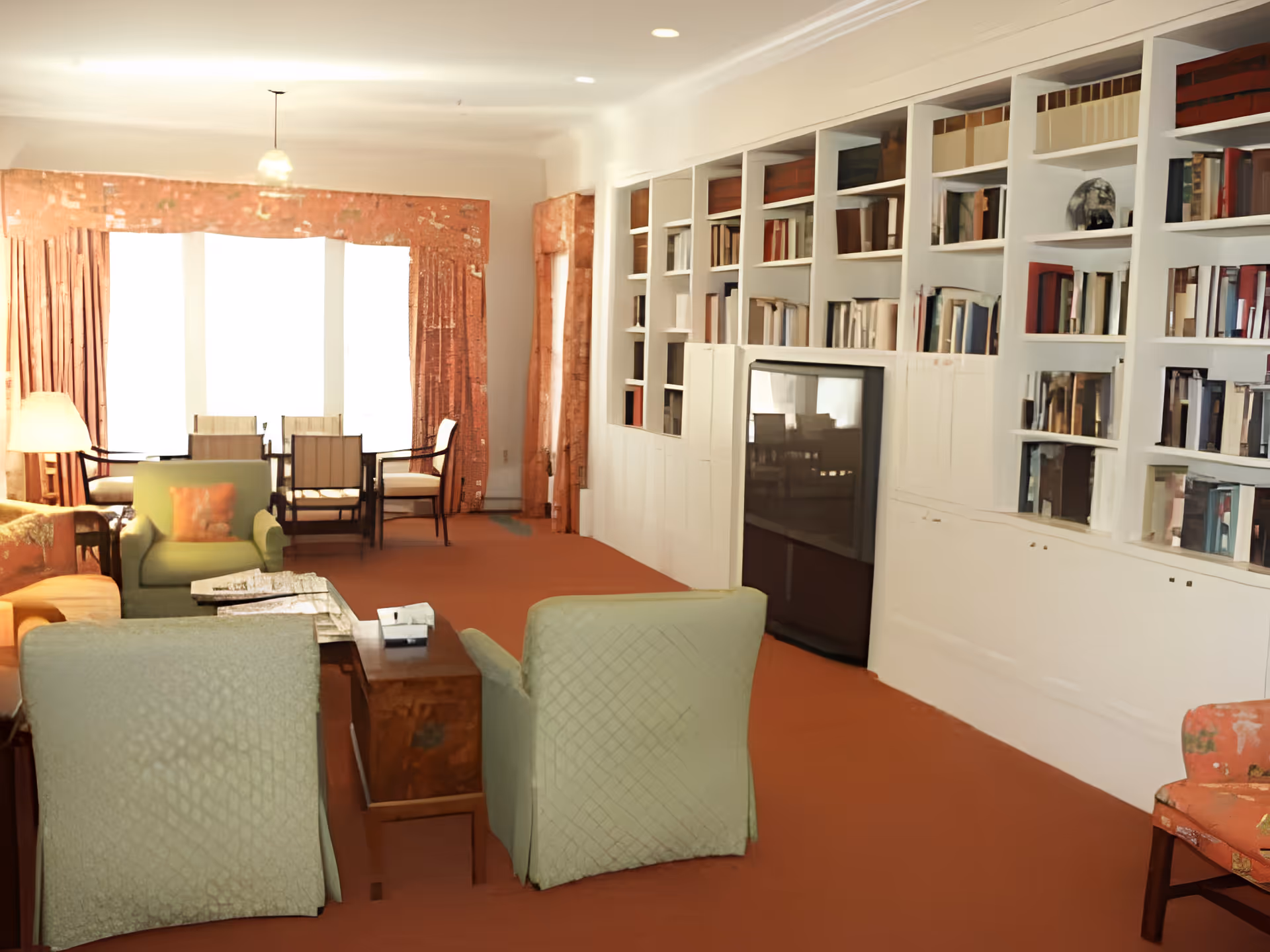 A cozy living room area with green armchairs, a wooden coffee table, and a large built-in white bookshelf filled with books. There is a dining table with chairs near large windows covered by orange curtains, and a lamp providing warm lighting.