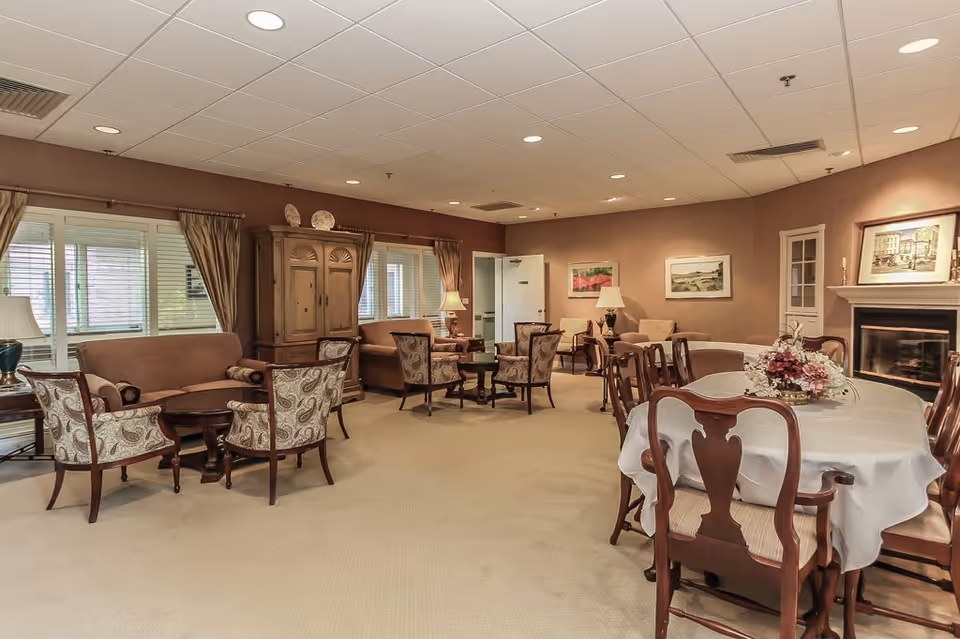 A spacious, warmly lit common area with multiple seating arrangements including upholstered armchairs, sofas, and wooden dining chairs around a table with a white tablecloth and floral centerpiece. The room features beige walls, carpeted floor, large windows with curtains, framed artwork, and a fireplace with a painting above it.