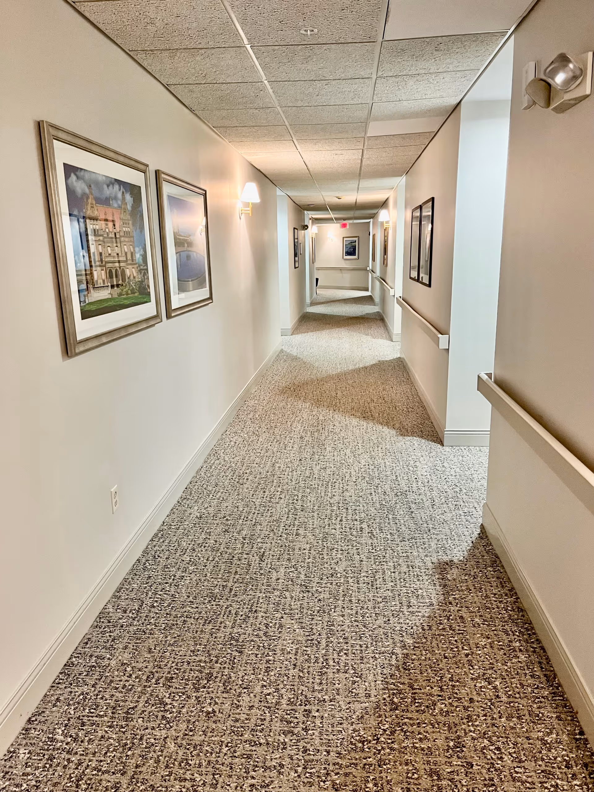 A long, carpeted hallway in a senior living facility with beige walls, handrails on both sides, framed pictures hanging on the walls, and ceiling lights providing illumination.