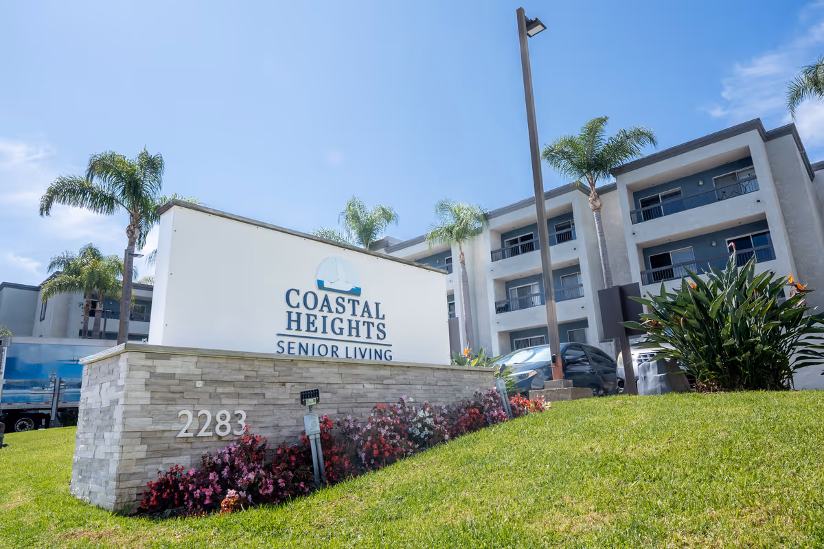 Exterior front of the Coastal Heights Senior Living building with its entrance sign, landscaped lawn, and palm trees.