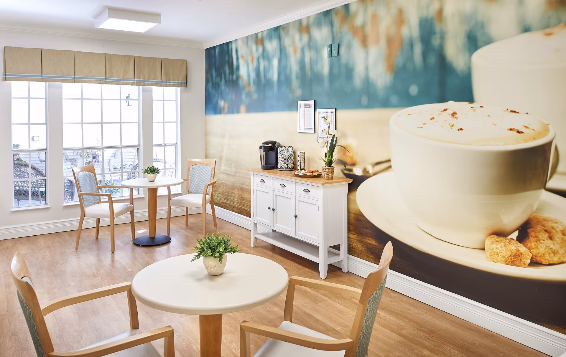 A bright and cozy dining area with wooden floors, two round tables each surrounded by two wooden chairs with light blue cushions. A white sideboard against the wall holds a coffee maker, cups, and a small plant. The wall behind the sideboard features a large mural of a coffee cup with frothy cappuccino and sugar cubes. Large windows with beige valances let in natural light, and there are small potted plants on the tables.