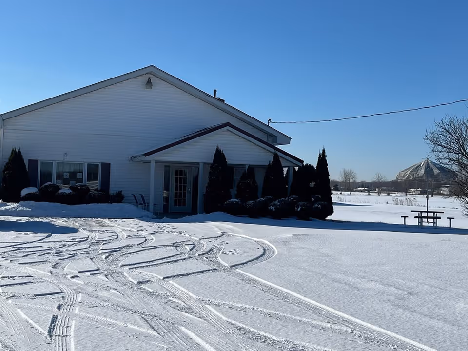 Exterior view of a white building with a covered entrance and several tall bushes in front. The ground is covered with snow, showing tire tracks. In the background, there is a picnic table and a large dome-shaped structure under a clear blue sky.