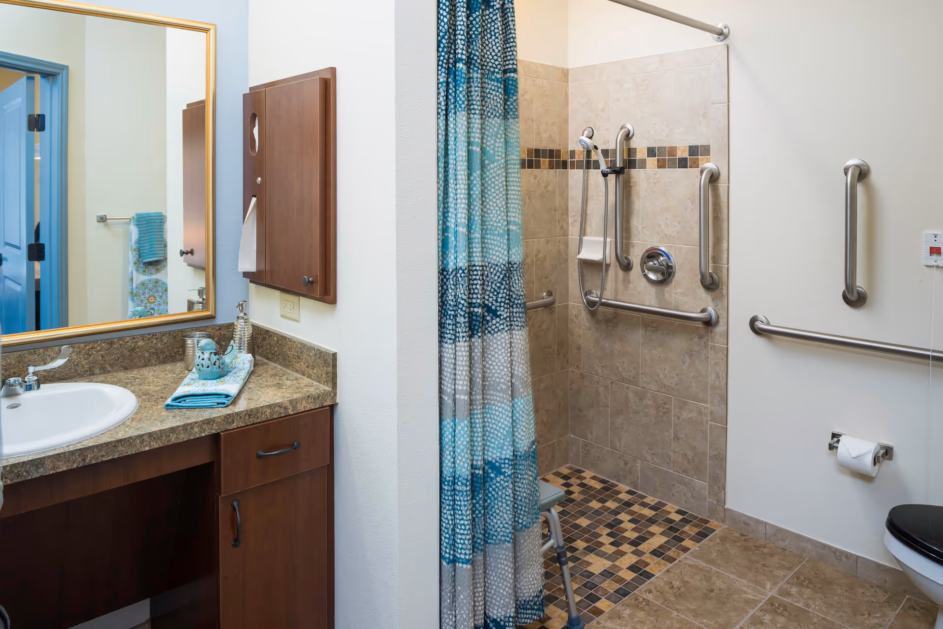 A bathroom with a sink and countertop on the left, a large mirror above the sink, and a wooden cabinet mounted on the wall. To the right is a walk-in shower with beige and brown tiled walls and floor, equipped with multiple grab bars and a handheld showerhead. A blue and white patterned shower curtain partially covers the shower entrance. There is a toilet with a black seat on the far right and a toilet paper holder mounted on the wall. The bathroom is designed for accessibility with grab bars and a shower chair visible inside the shower.