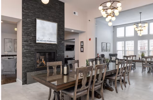 A spacious and well-lit dining area with multiple wooden tables and chairs arranged neatly. The room features a modern black stone fireplace with a framed painting above it. Large windows with white blinds allow natural light to fill the space. Contemporary chandeliers hang from the ceiling, and there are decorative plants and framed artwork on the walls.