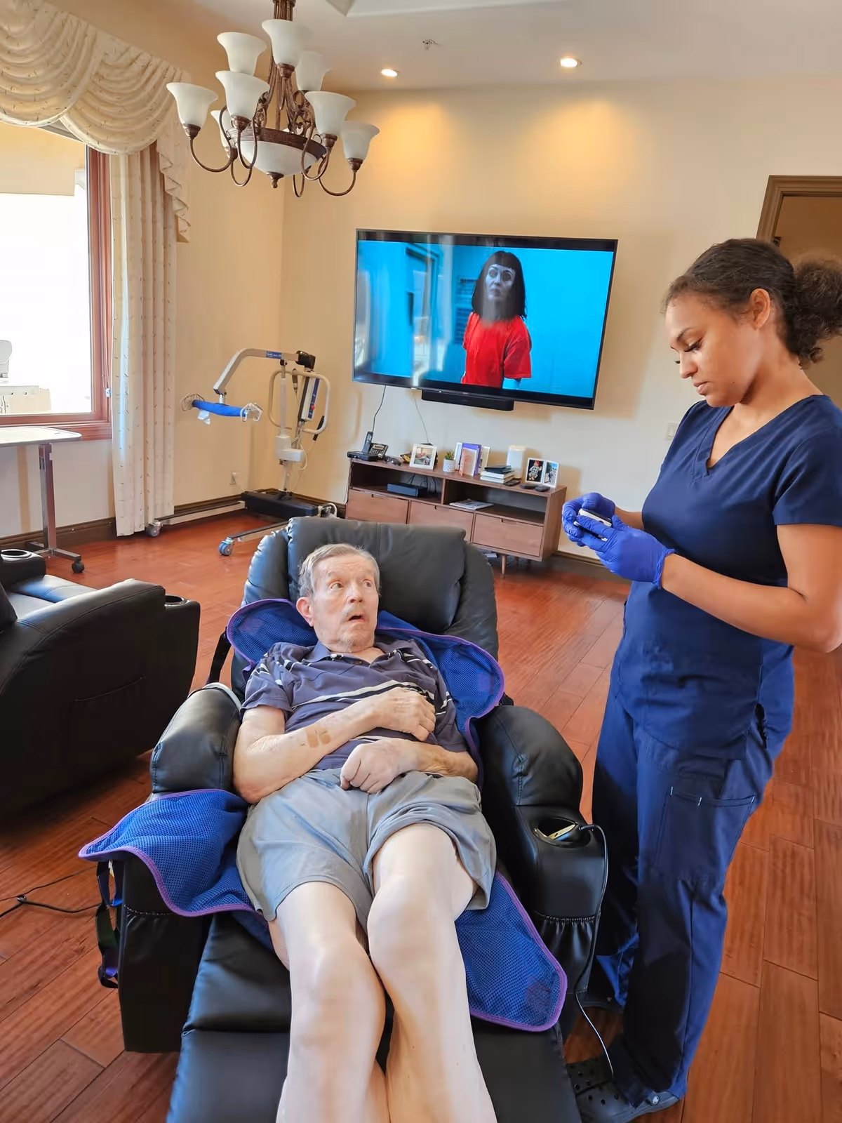 An elderly man reclining in a black leather chair covered with a blue blanket in a living room. A caregiver wearing navy blue scrubs and purple gloves stands beside him, preparing to assist. The room has wooden flooring, a large window with curtains, a chandelier, a TV mounted on the wall displaying a woman in a red shirt, and a wooden cabinet with framed photos and other items.