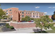 Brick multi-story senior healthcare building with a heart logo, raised brick planter and landscaping in front under a blue sky.