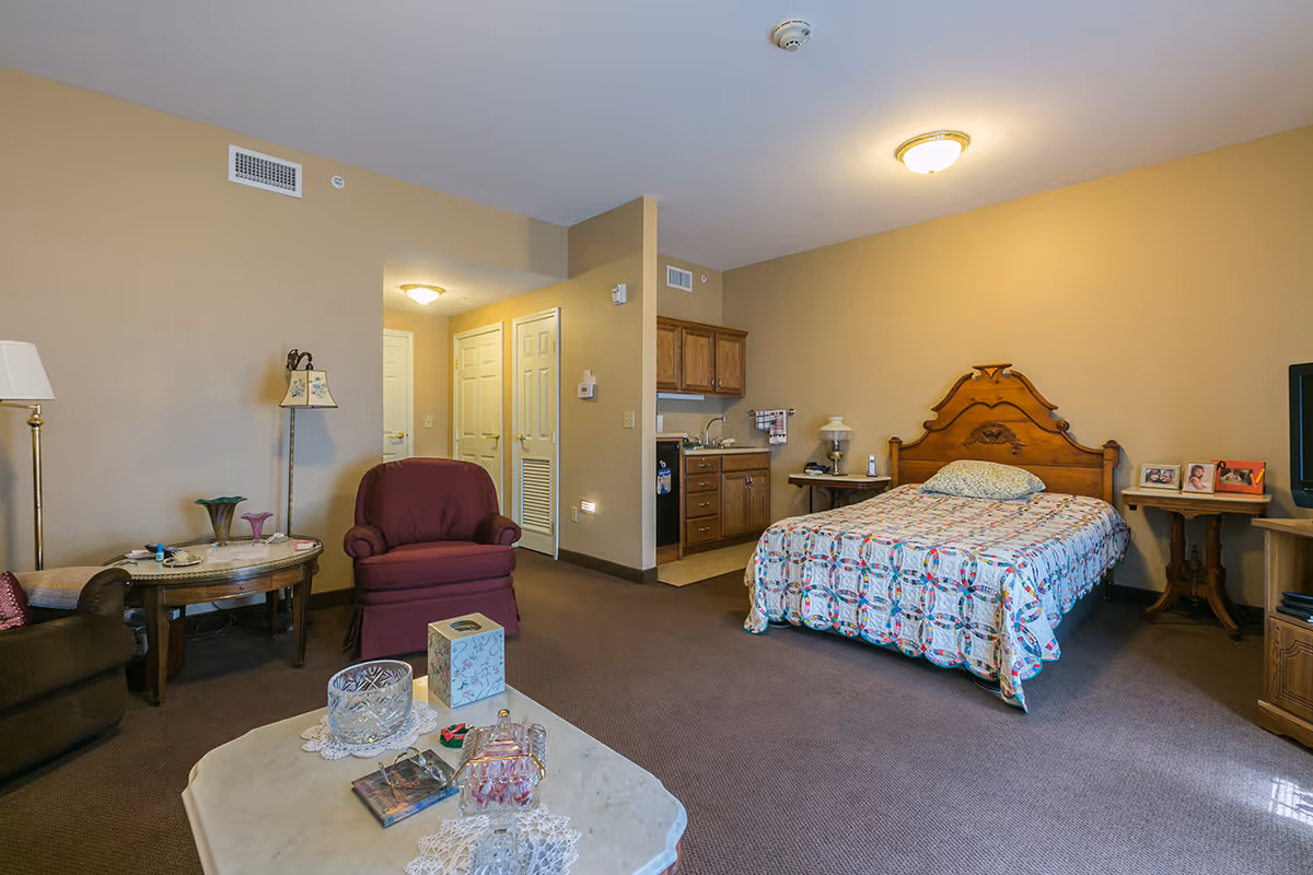 Interior view of a senior living facility room at The Bungalows at Fayetteville, featuring a bed with a patterned quilt, a small kitchenette with wooden cabinets, a maroon armchair, a brown armchair, a coffee table with decorative items, and beige walls with carpeted floor.