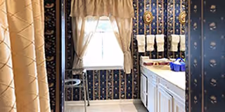 A bathroom with dark blue floral wallpaper, a white countertop with a sink, several white towels hanging on a rack, a window with white curtains, and a beige shower curtain partially visible on the left side.