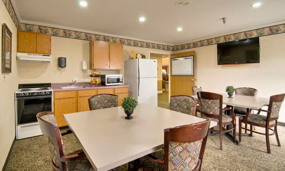 A communal dining and kitchen area with two square tables surrounded by patterned chairs. The kitchen area includes a stove, sink, microwave, refrigerator, and wooden cabinets. A flat-screen TV is mounted on the wall, and a small potted plant is on each table. The room has patterned carpet and wallpaper border near the ceiling.