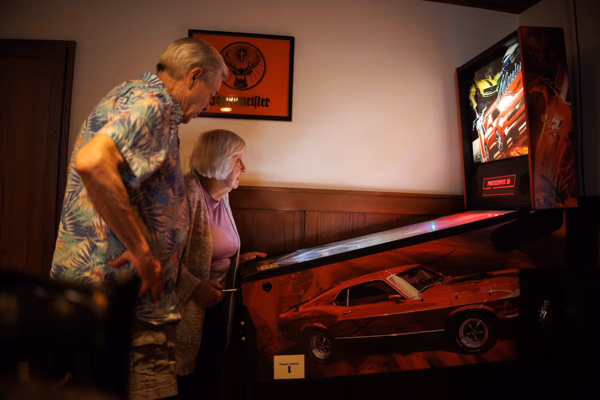 An elderly man and woman playing a red pinball machine with a car design on its side in a room with wood paneling and a framed Jägermeister poster on the wall.