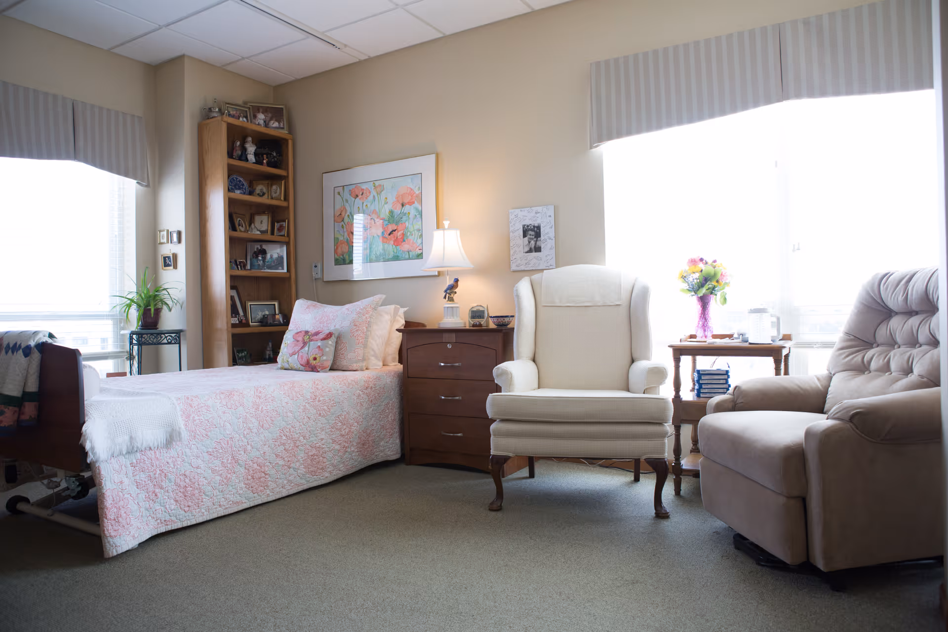 A cozy bedroom in a senior living facility with a single bed covered in a pink and white quilt, a wooden nightstand with a lamp, a cushioned armchair, a recliner, a wooden bookshelf filled with framed photos and decorations, and a small table with a vase of flowers near a large window with striped valances.