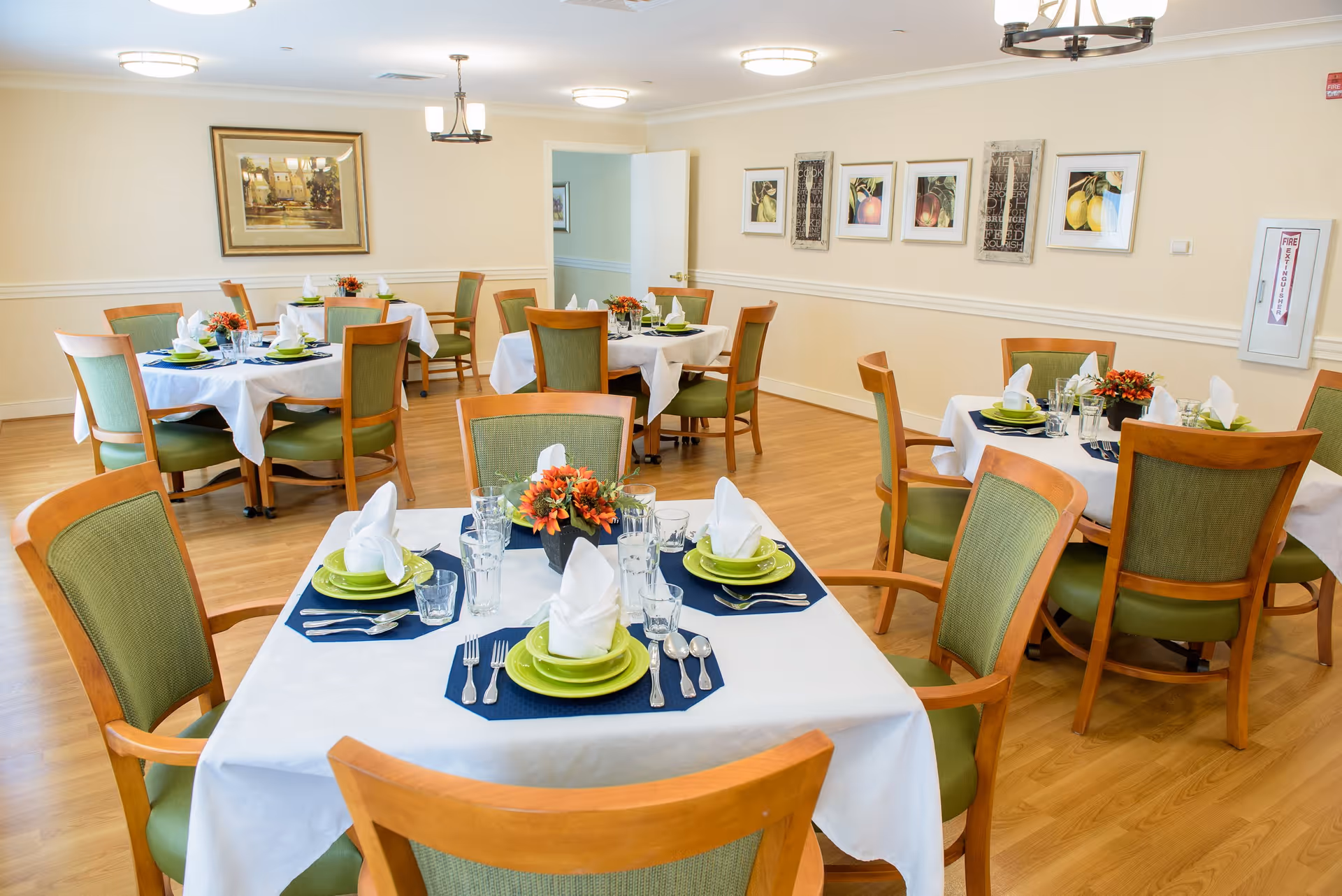 A bright dining room with several round and square tables set with white tablecloths, green plates, folded white napkins, glassware, and silverware. Each table has a small floral centerpiece. The room has wooden floors, green cushioned wooden chairs, framed artwork on the walls, and ceiling light fixtures.