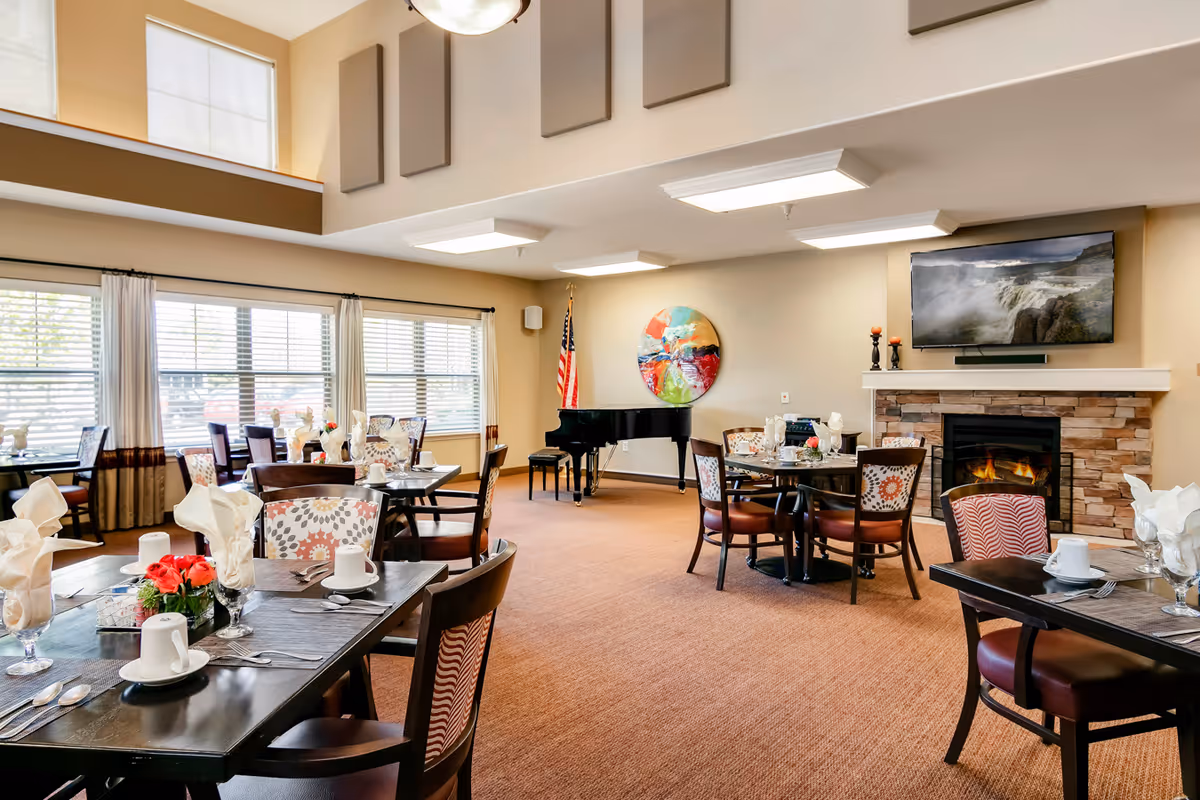 A bright and spacious dining room with several tables set with white napkins, cups, and silverware. The room features large windows with blinds and curtains, a fireplace with a TV mounted above it, a colorful round wall art, and a black grand piano near an American flag.