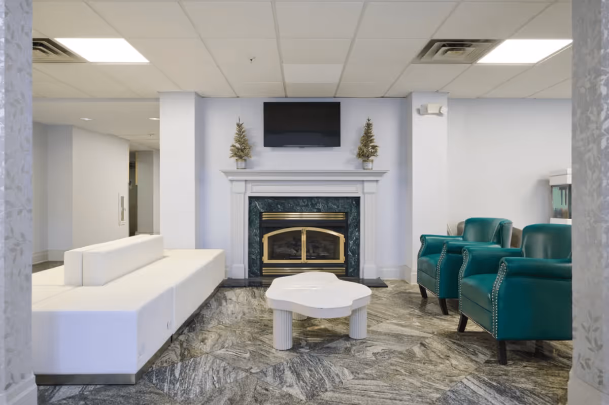 A modern living room area with a white sectional sofa on the left, two green armchairs on the right, a white coffee table in the center, and a fireplace with a dark green marble surround and brass accents. Above the fireplace is a flat-screen TV mounted on the wall, flanked by two small decorative plants. The floor has a gray and white patterned tile, and the ceiling has recessed lighting.