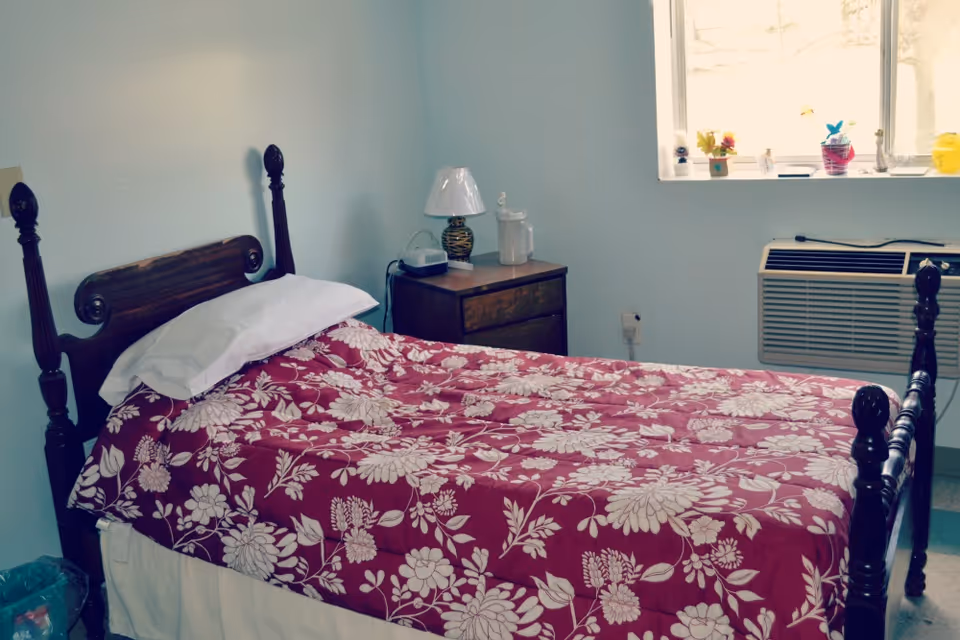 Single bed with a red floral bedspread, wooden headboard, nightstand with a lamp, a window, and a wall air conditioner.