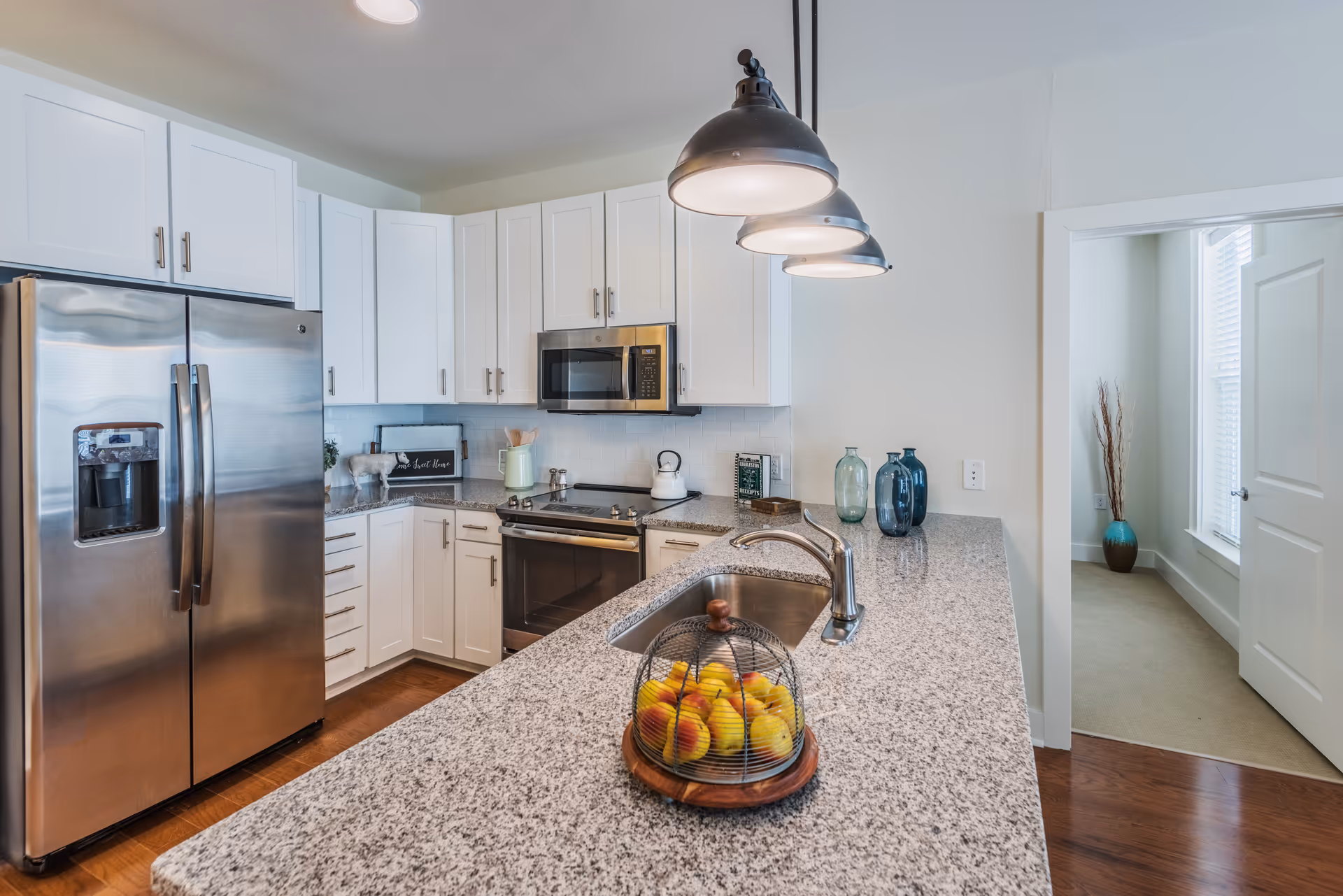 Modern kitchen with white cabinets, stainless steel refrigerator, microwave, and oven. Granite countertop island with a sink and a decorative fruit basket containing apples. Three pendant lights hang above the island. Adjacent room with a large window and a decorative vase with tall branches is visible through an open doorway.