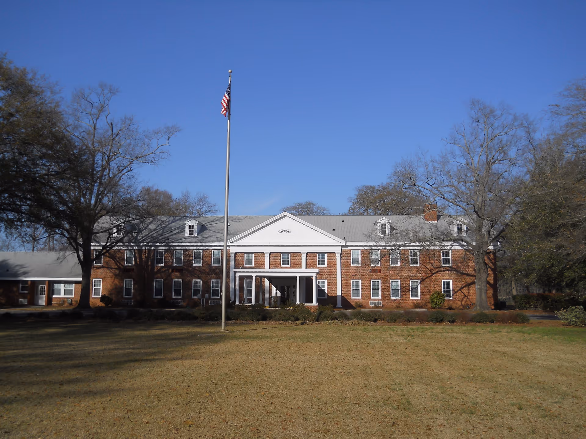 Front exterior view of a large two-story brick building with white trim and a gray roof, featuring multiple windows and a central entrance with columns. An American flag on a tall flagpole stands in front of the building, surrounded by trees and a grassy lawn under a clear blue sky.
