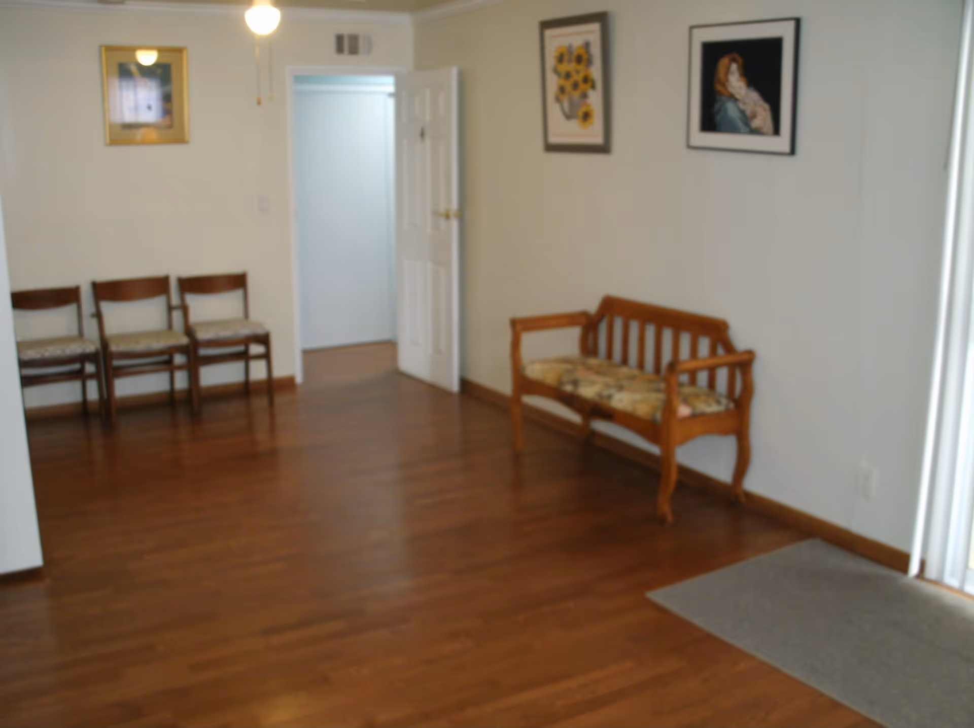 A room with wooden flooring featuring a wooden bench with a floral cushion on the right wall and three wooden chairs with cushions lined up against the left wall. There are two framed pictures hanging on the right wall above the bench and one framed picture on the left wall. A white door is open in the center background, and a ceiling fan with a light is visible on the ceiling.