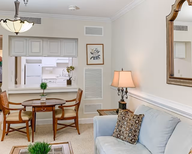 A cozy living area with a light blue sofa adorned with a patterned cushion, a wooden side table with a decorative lamp, and a wooden dining table with two chairs. The room has beige walls and carpet, a large mirror on the wall, and a pass-through window showing a white kitchen with cabinets, a refrigerator, and a sink.