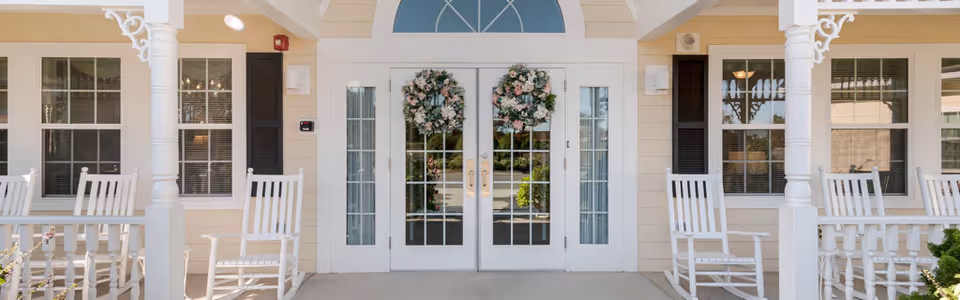 Front entrance of a senior living facility with double glass doors adorned with floral wreaths, white columns and porch rocking chairs.
