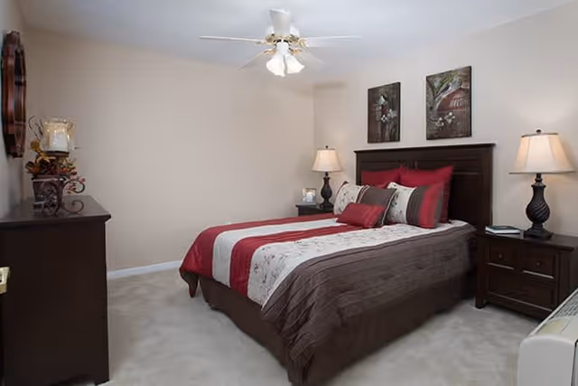 A neatly made bed with a brown, red, and white striped comforter and multiple pillows in a bedroom. The room has beige walls, a ceiling fan with lights, two bedside tables with lamps, a dresser with decorative items, and two framed pictures hanging above the bed.
