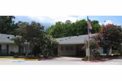 Front exterior view of College Street Health Care Center building with a covered entrance, American flag on a flagpole, trees, and landscaped bushes.