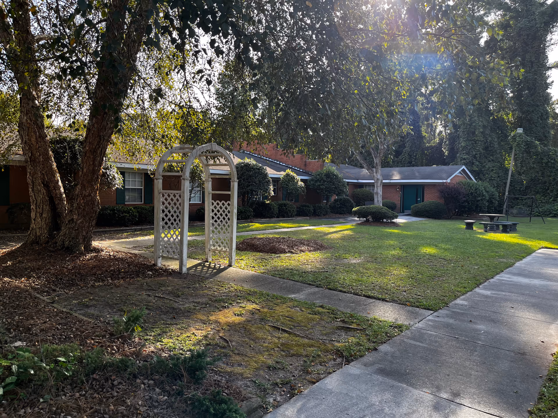 Outdoor courtyard area of a senior living facility with a white wooden archway, large trees, green grass, bushes, a concrete walkway, and a brick building with green doors and windows in the background. There is also a picnic table and benches on the right side.