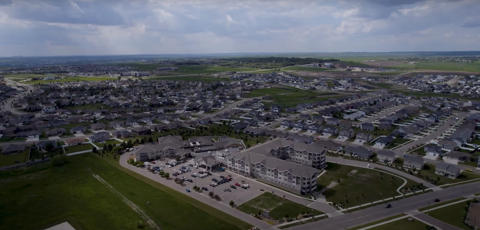 Aerial view of a large senior living facility named Benedictine Living Community-Bismarck surrounded by a residential neighborhood with many houses and green fields under a partly cloudy sky.