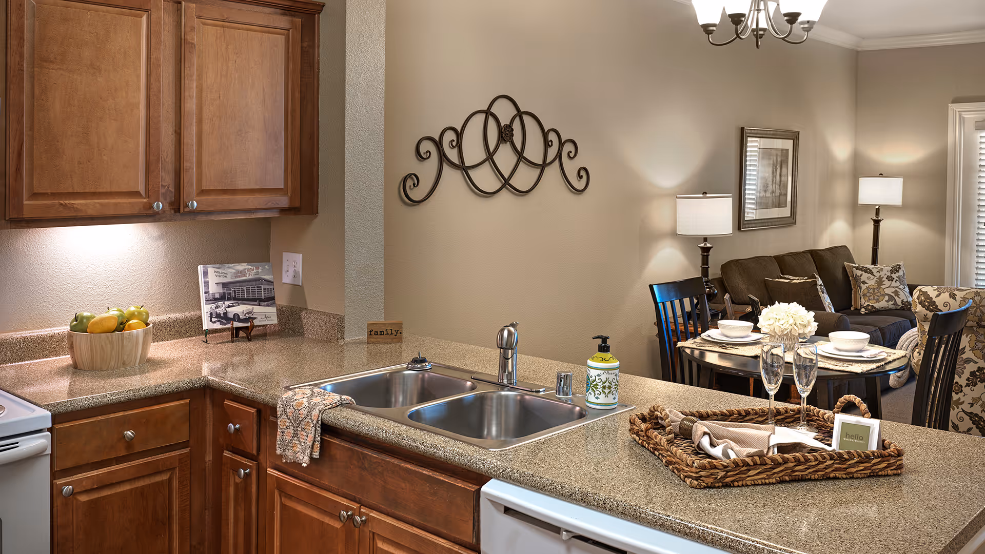Interior view of a kitchen and living room area in a senior living facility. The kitchen features wooden cabinets, a double sink, a countertop with a basket holding napkins and two empty champagne glasses, and a bowl of fruit. The living room has a dining table set with plates and bowls, a sofa with cushions, a floor lamp, and wall decorations including a framed picture and a decorative metal wall art piece.
