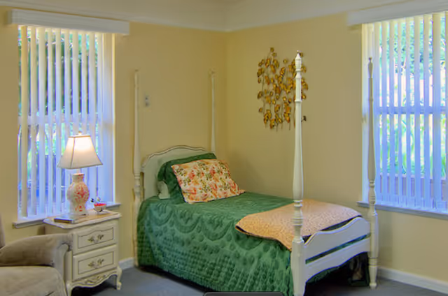 A bright bedroom featuring a white four-poster twin bed with a green quilt, a nightstand with a lamp, and two tall windows with vertical blinds.