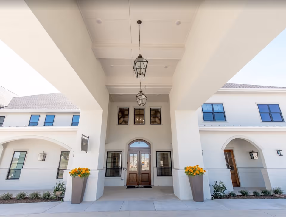 Entrance of a senior living facility with a covered driveway, two large planters with yellow flowers on either side, white walls, wooden double doors, and hanging lantern-style light fixtures.