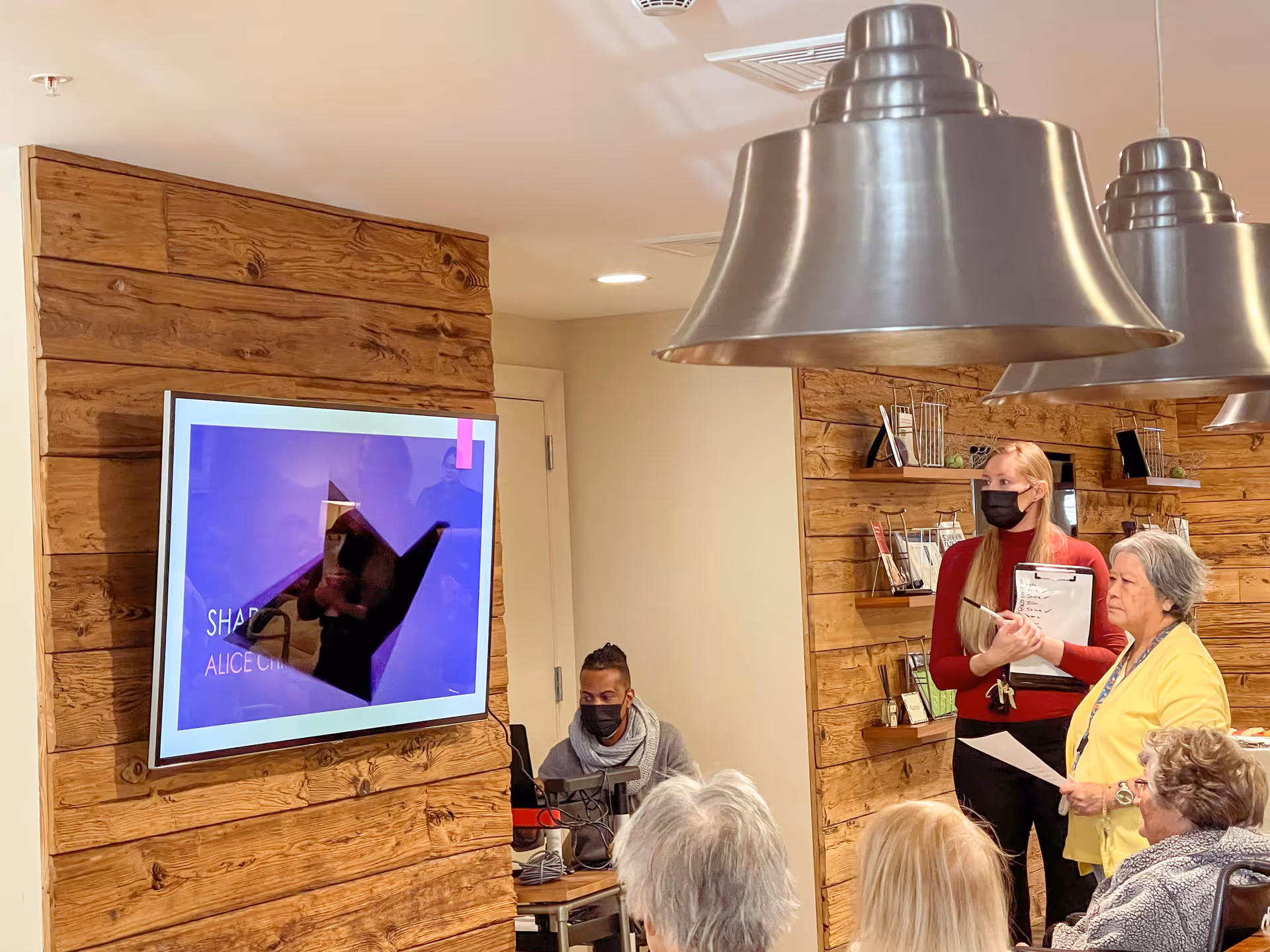 A group of elderly people seated and watching a presentation on a wall-mounted TV screen in a room with wooden panel walls. A woman in a red sweater and black mask is standing and holding a clipboard, while another woman in a yellow sweater is holding a paper. The room has modern hanging metal light fixtures.
