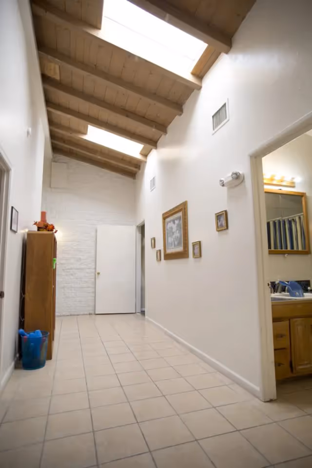 A bright hallway with a tiled floor and a wooden ceiling featuring skylights. The walls are white with several framed pictures hanging on the right side. On the left side, there is a wooden cabinet with some decorative items on top and a blue trash bin on the floor. An open door on the right reveals a bathroom with a sink and mirror.