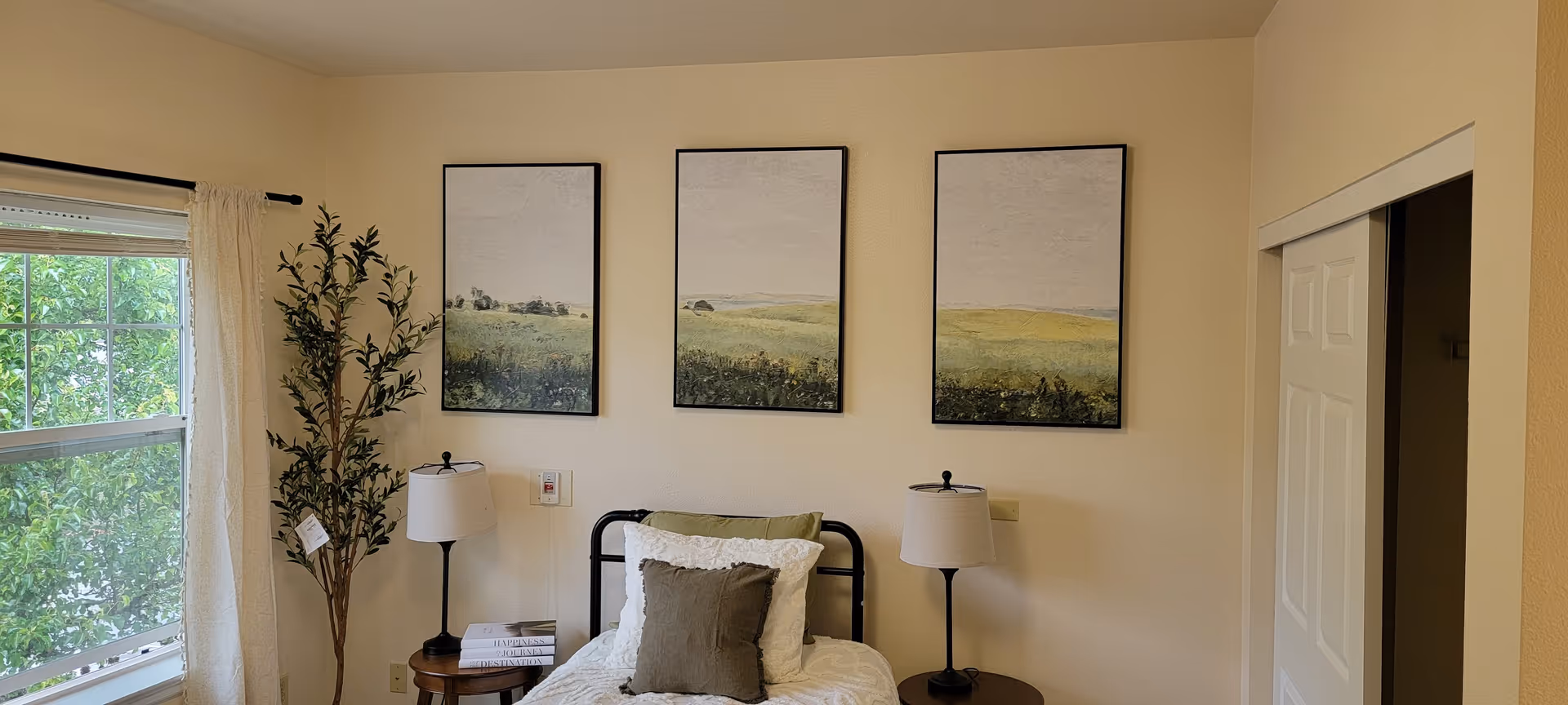 A bedroom with a single bed featuring white bedding and a gray pillow. On either side of the bed are small round wooden tables with matching lamps. Above the bed are three framed landscape paintings. To the left, there is a window with sheer curtains and a tall potted plant beside it. On the right side, there is a white sliding closet door.