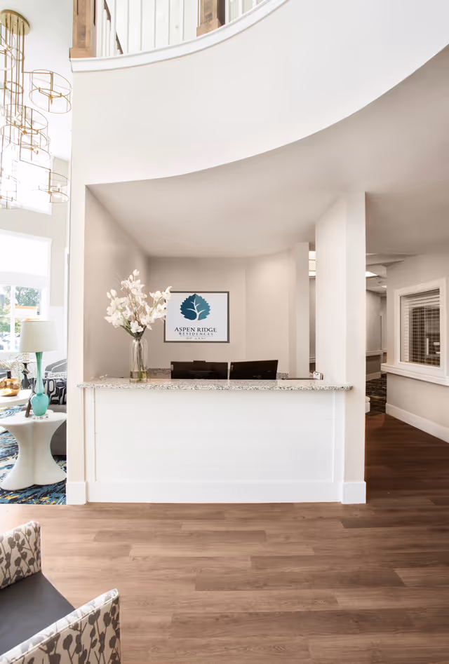 Reception desk area inside Aspen Ridge Residences of Lehi with a granite countertop, a vase with white flowers, two computer monitors, and a framed Aspen Ridge Residences logo on the wall behind. The space has light-colored walls, wood flooring, and partial views of seating areas and decorative lighting.