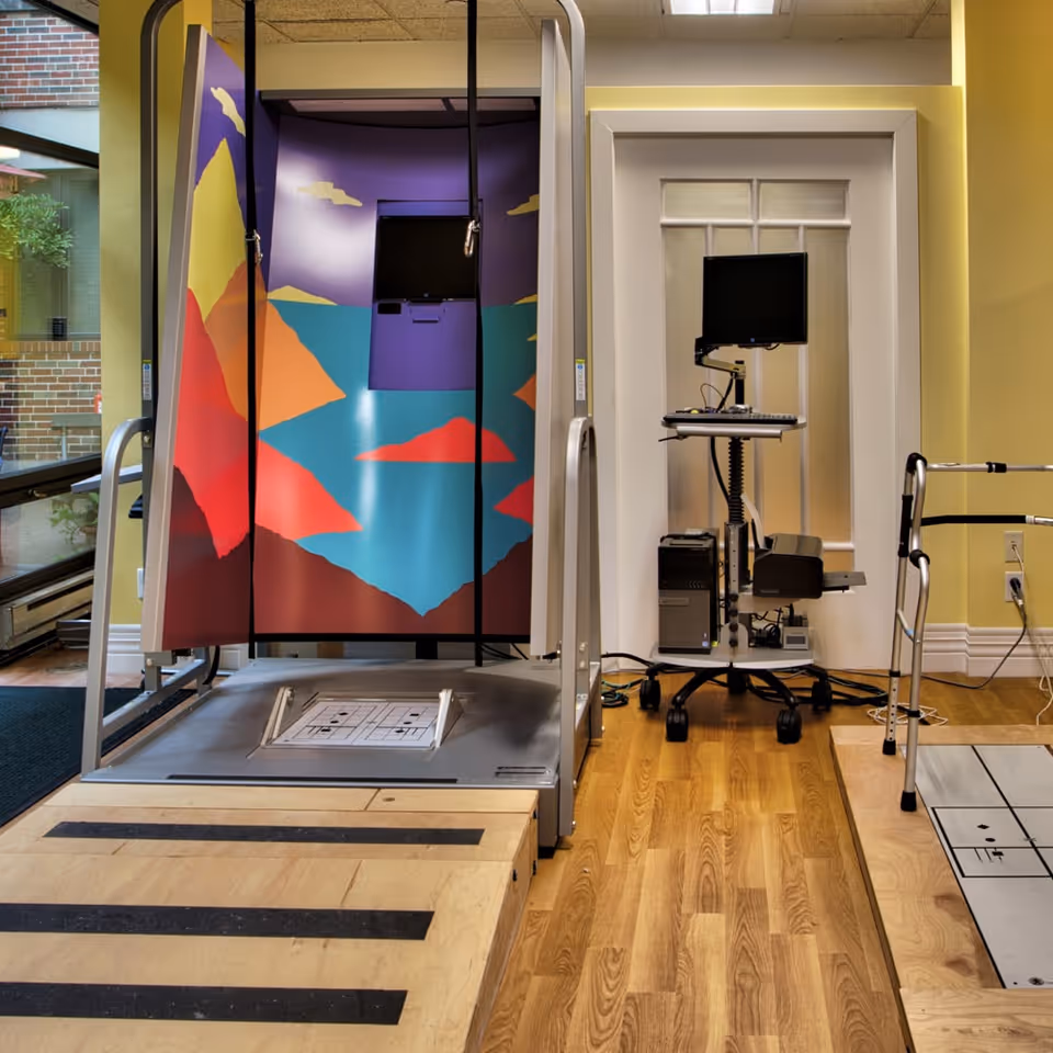 A rehabilitation or physical therapy room with a colorful enclosed treadmill on the left, a computer workstation with a monitor and keyboard on a mobile stand in the center, and a walker on the right. The room has wooden flooring and a large window showing an outdoor brick wall and greenery.