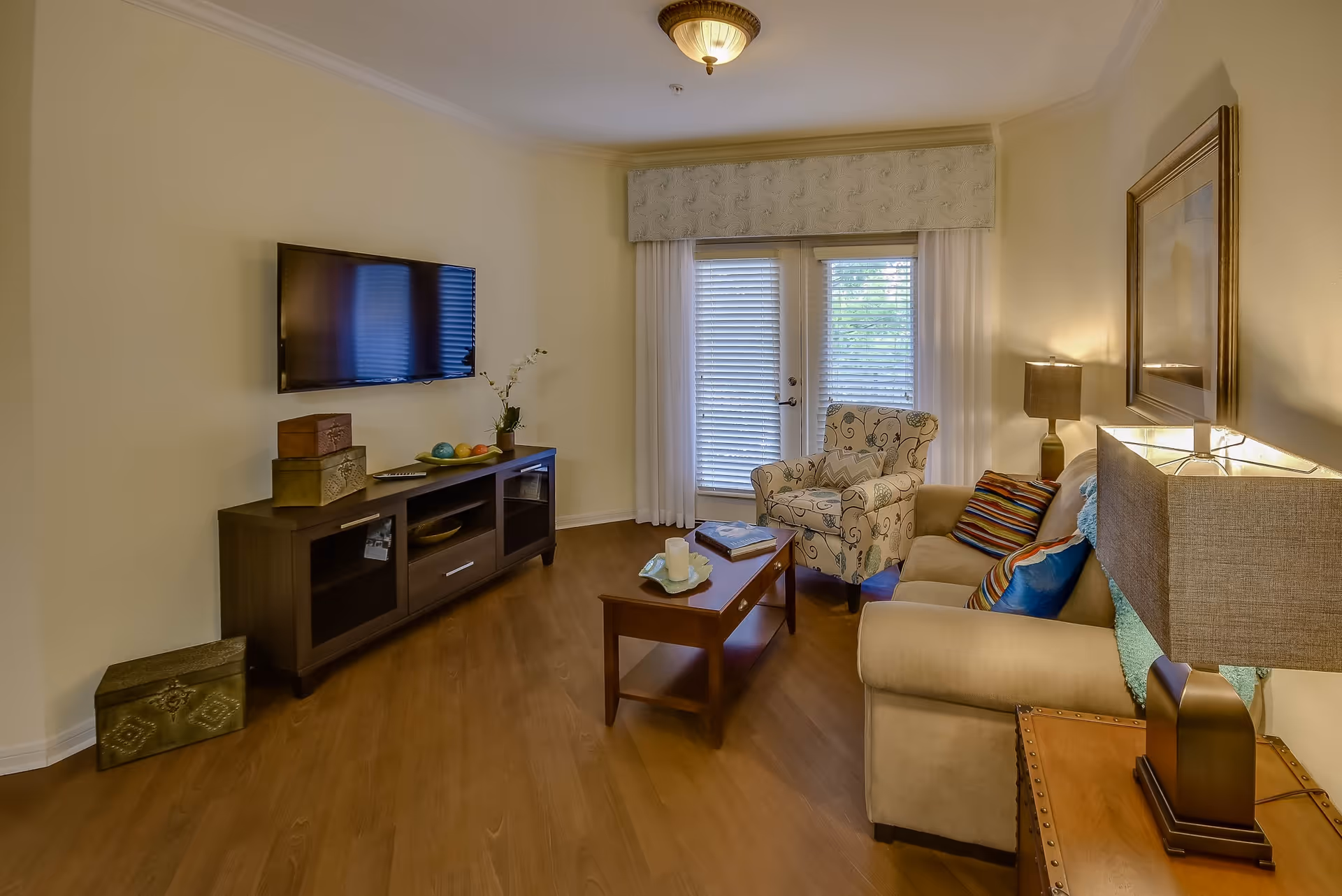 A cozy living room with a beige sofa adorned with colorful pillows, a patterned armchair, a wooden coffee table with candles and a book, a TV mounted on the wall above a dark wooden media console, and a side table with a lamp. French doors with blinds and curtains lead outside, and the room has warm wooden flooring and soft yellow walls.