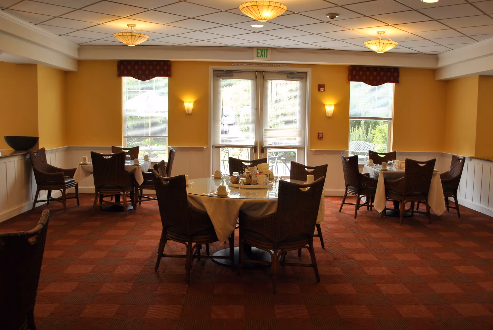 A dining room with round tables covered with white tablecloths and set with cups and condiments. The room has yellow walls, large windows with valances, wall sconces, and a glass door leading outside. The floor is carpeted in a red and brown checkered pattern, and wicker chairs surround the tables.