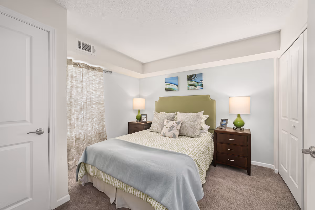 A tidy bedroom featuring a bed with a green headboard, bedside tables with lamps, artwork above the bed, and a window with curtains.