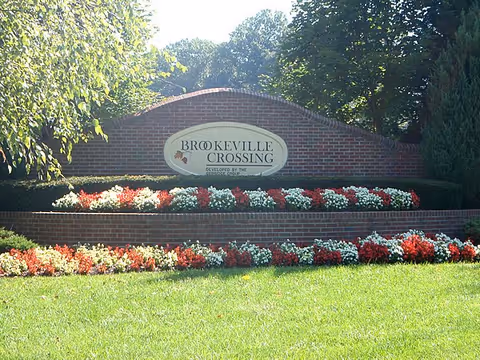 Brick entrance sign reads Brookeville Crossing with red and white flowers and a green lawn.