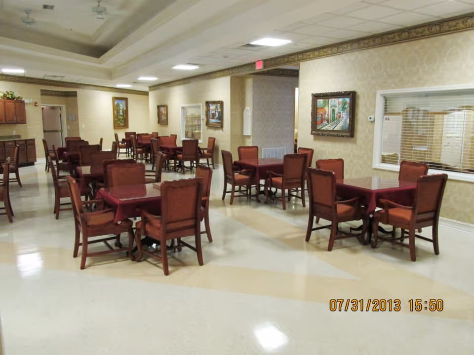 A dining room with multiple square tables covered with burgundy tablecloths, each surrounded by four wooden chairs with red upholstery. The room has beige patterned wallpaper, framed paintings on the walls, and a window with blinds looking into another room. The floor is a light-colored polished surface, and the ceiling has recessed lighting and a decorative border near the top of the walls.