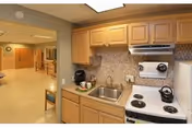 Interior view of a small kitchenette area with wooden cabinets, a stainless steel sink, a white electric stove with four burners, a kettle, and a coffee maker. The kitchenette opens into a hallway with beige walls and flooring, and a wooden chair is partially visible.