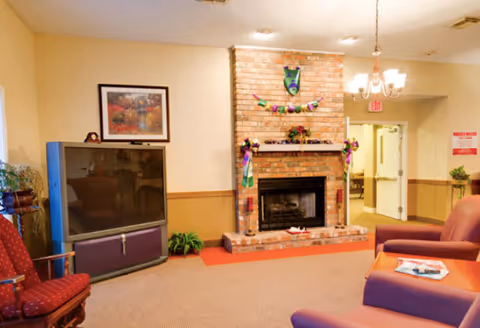 A cozy living room area with a brick fireplace decorated with colorful Mardi Gras-themed garlands and masks. There are comfortable chairs and a large television on a stand to the left. The walls are painted beige with a wooden chair rail, and a framed picture hangs above the television. A chandelier light fixture hangs from the ceiling, and an open doorway leads to a hallway.