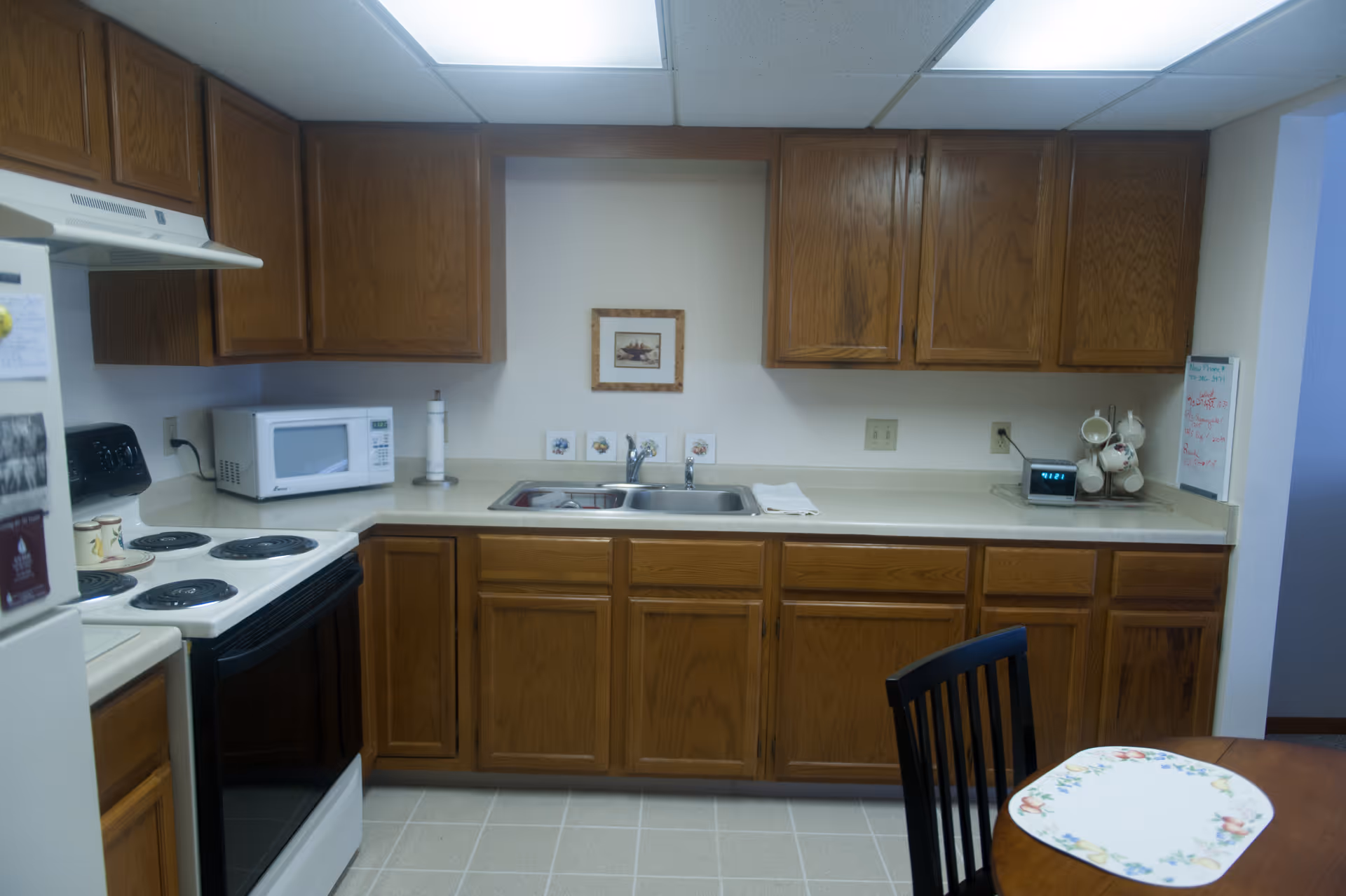 A kitchen with wooden cabinets, a white electric stove, a microwave, a double sink, and a small round dining table with a floral placemat and a black chair. The countertop has a paper towel holder, a clock, and a mug rack with cups. There is a small framed picture on the wall above the sink.