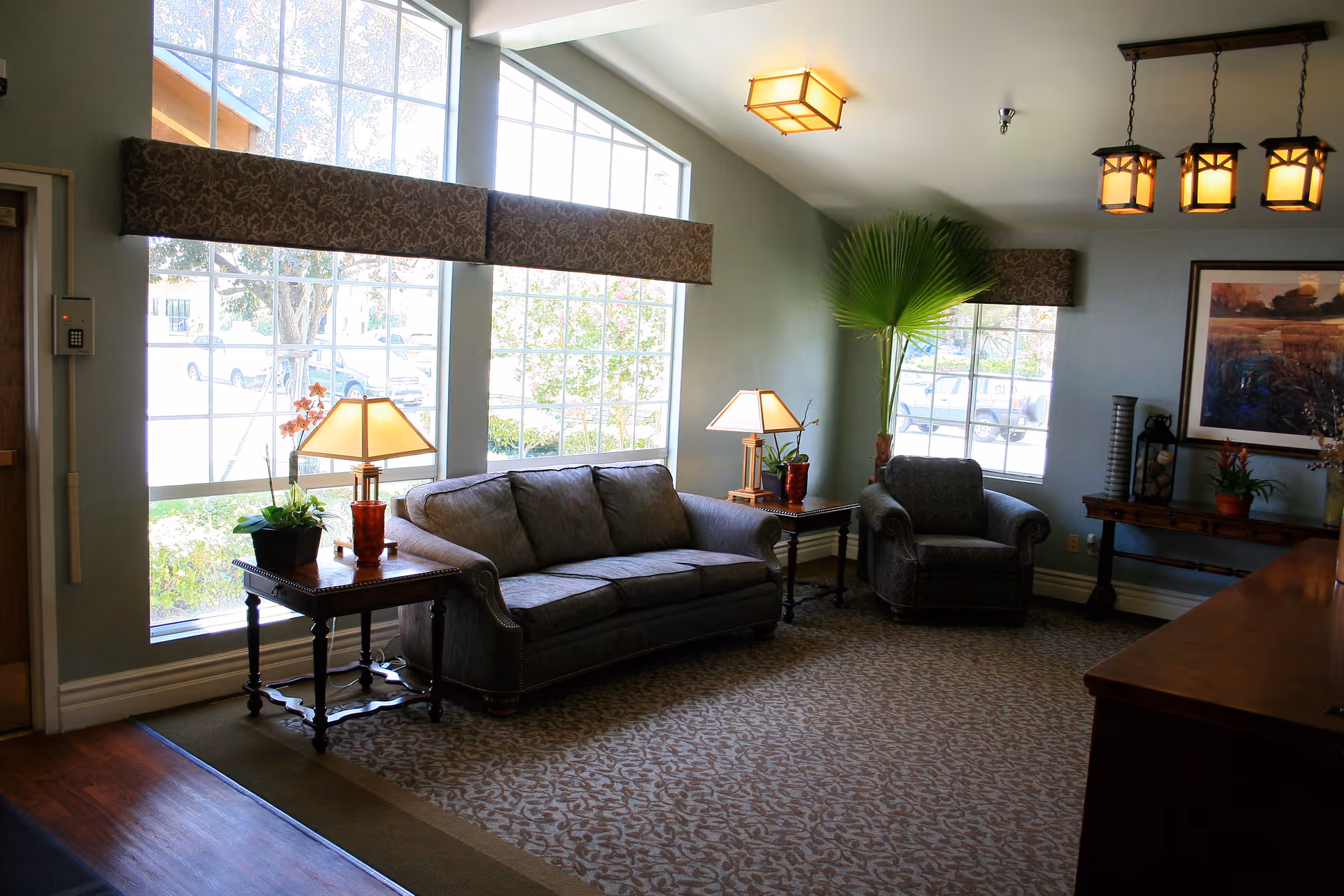 A cozy living room area with a gray sofa and matching armchair, two wooden side tables with lamps, large windows letting in natural light, a patterned carpet, a large green plant in the corner, and a framed painting on the wall.