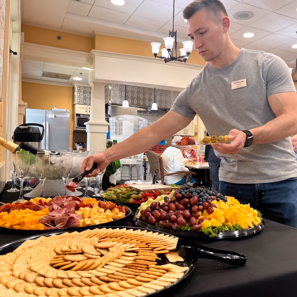 A man serving himself from a buffet table with platters of crackers, cheese, fruit and charcuterie in a dining area.