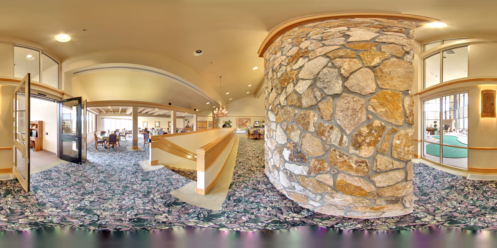 Spacious senior living common area with a large stone pillar, patterned carpet, and tables and seating under a vaulted ceiling.