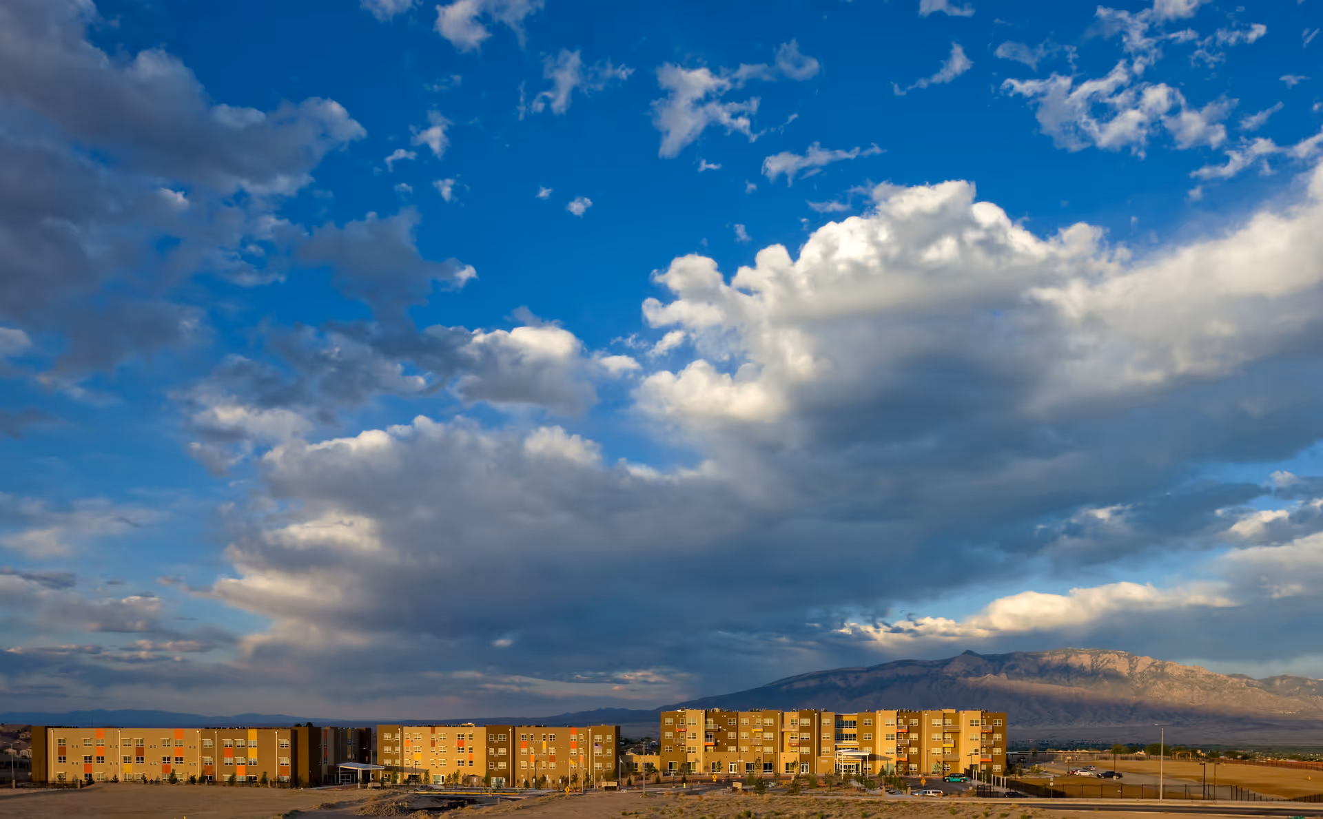 Low-rise apartment-style senior living complex set under a vast blue sky with dramatic clouds and distant mountains.