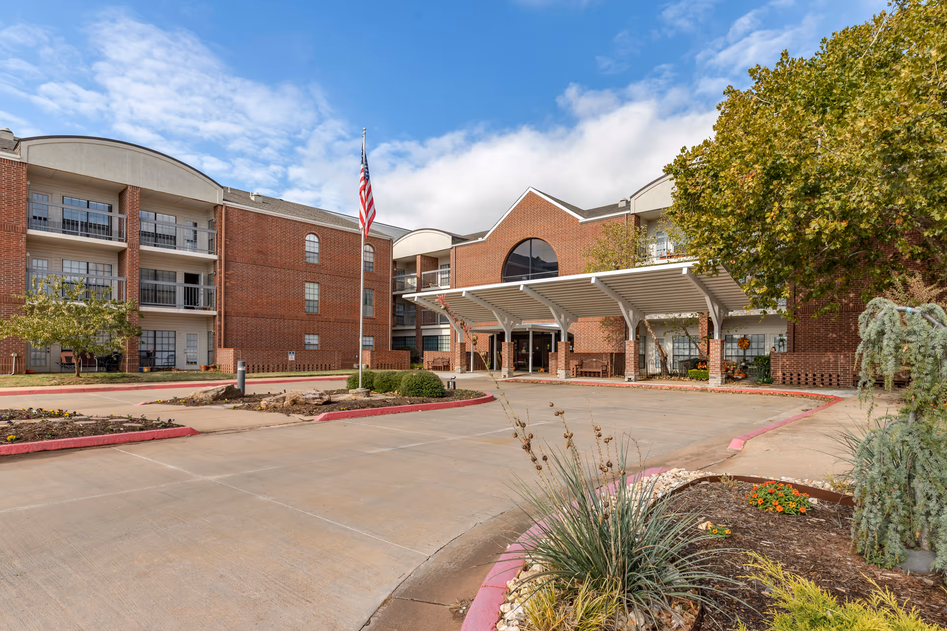 Exterior view of a senior living facility with a brick facade, multiple floors with balconies, an American flag on a flagpole, a covered entrance, and landscaped areas with trees and plants under a partly cloudy sky.